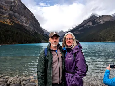 A photograph of a middle-aged male and female, with their arms linked, standing on the shore of a large deep-blue coloured lake. The couple are facing the camera, and the lake fills the background.