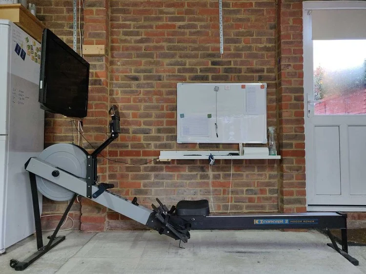 A side-on photograph of a Concept 2 rowing maching in front of a wall and a wall mounted TV.
